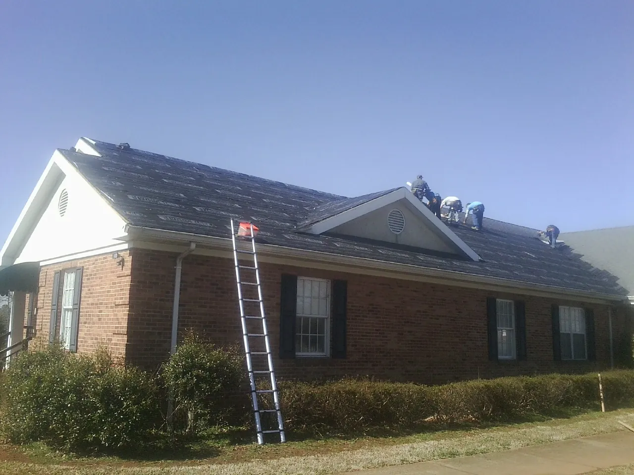 Crew tearing shingles off a brick ranch roof with a ladder set — Emergency Roof Repair work by our crew in Holgate